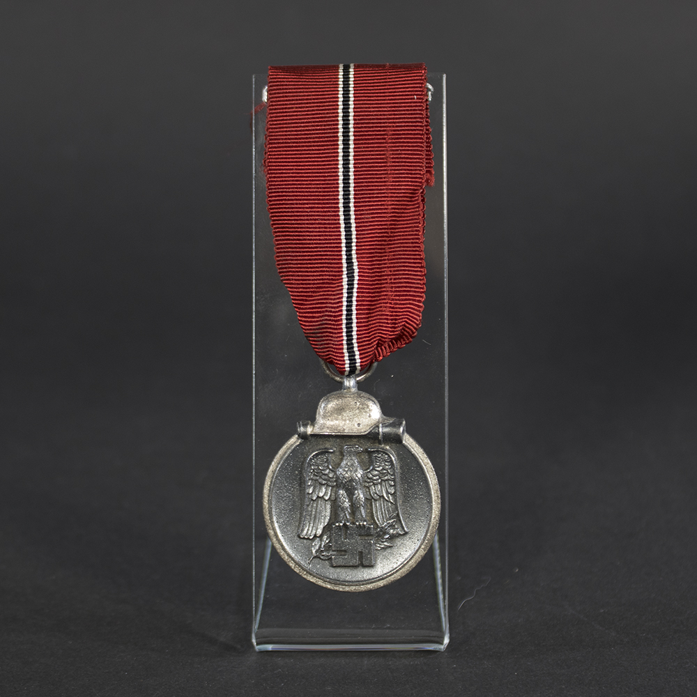 Front of a German Eastern Front Medal showing a raised design with a German eagle clutching a swastika above a crossed sword and laurel wreath, with a helmet and grenade motif at the bottom.