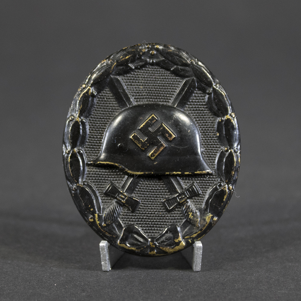 Front of a black wound badge showing an embossed German helmet with a swastika in the center, crossed swords behind it, and a textured oval background surrounded by a laurel wreath.