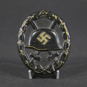 Front of a black wound badge showing an embossed German helmet with a swastika in the center, crossed swords behind it, and a textured oval background surrounded by a laurel wreath.