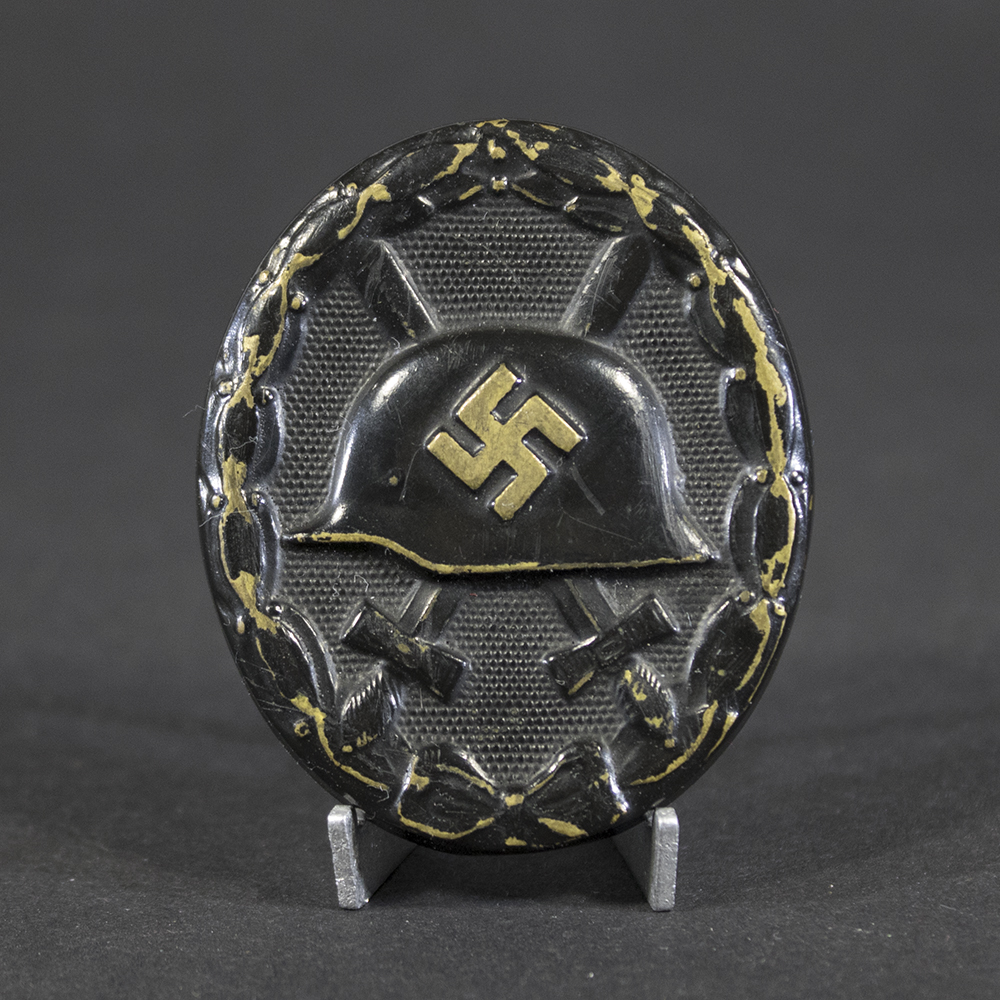 Front of a black wound badge showing an embossed German helmet with a swastika in the center, crossed swords behind it, and a textured oval background surrounded by a laurel wreath.