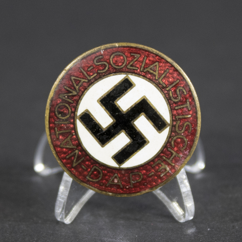 Front of an NSDAP membership badge showing a circular design with a white enamel ring inscribed ‘National-Sozialistische D.A.P.’ around a red background featuring a black swastika.