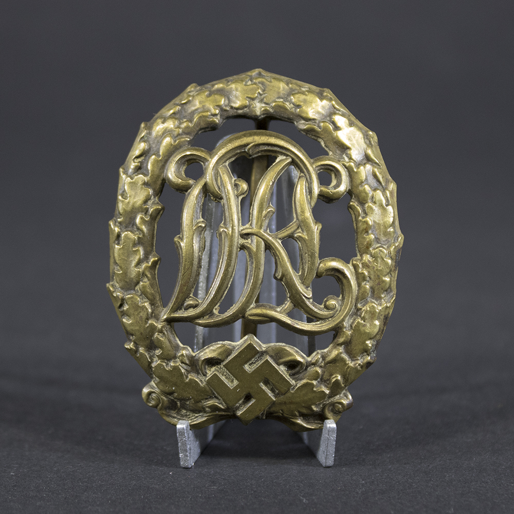 Front of a DRL Sport Badge featuring an ornate monogram of the letters ‘DRL’ in a decorative script, surrounded by an oval laurel wreath with a swastika at the bottom.