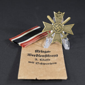 A WWII German War Merit Cross with swords displayed next to its original light brown paper bag. The bronze-toned medal hangs from a ribbon, and the bag shows printed German text.