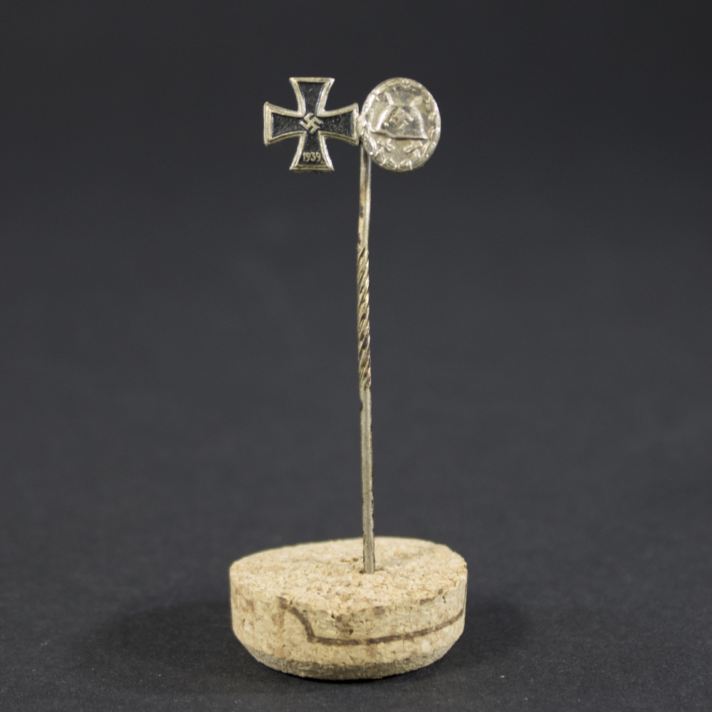 A WWII German stick pin featuring a miniature Iron Cross and a silver wound badge mounted on a single pin. Both medals are finely detailed with a metallic finish.
