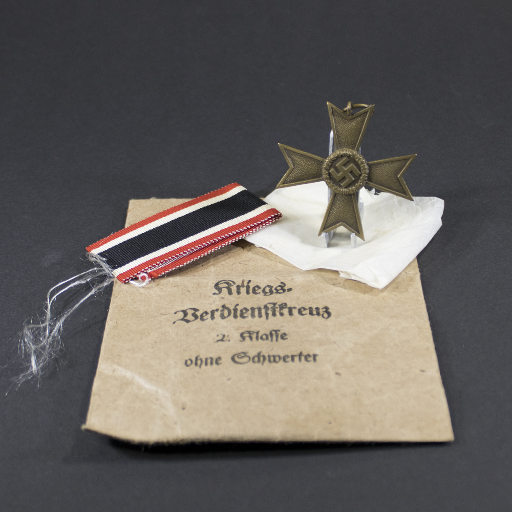 A WWII German War Merit Cross without swords displayed next to its original light brown paper bag. The bronze-toned medal hangs from a ribbon, and the bag shows printed German text.