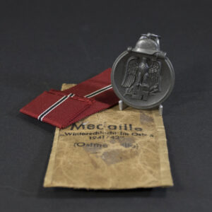 A WWII German Eastern Front Medal placed next to its original paper bag. The medal is dark gray with a loop at the top, and the bag is light brown with printed German text.