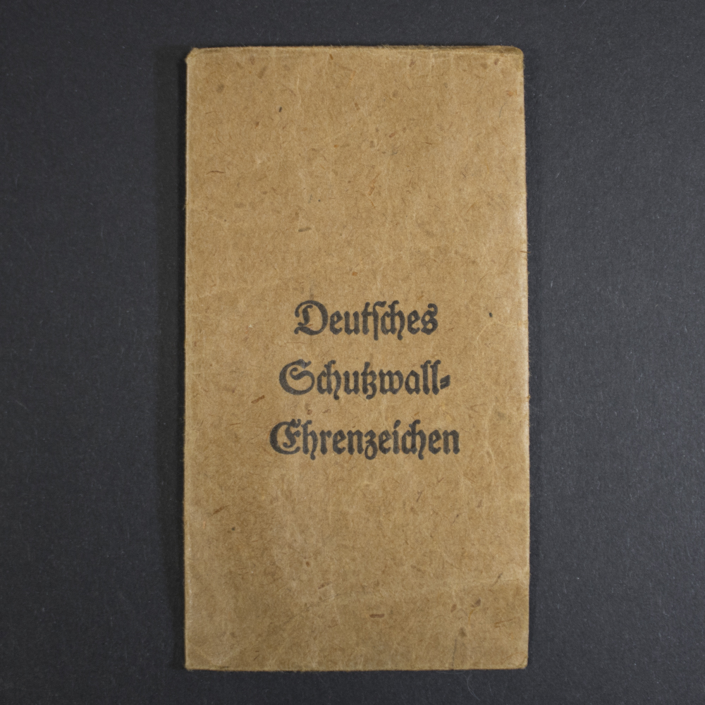 Light brown paper bag with printed text “Deutsches Schutzwall-Ehrenzeichen” in black ink. The surface shows creases and aging spots.