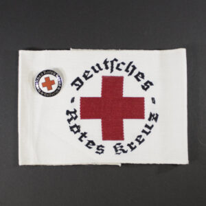 “German Red Cross (DRK) armband displayed together with a Helferin badge. The armband is white with a prominent red cross in the center, while the badge is oval-shaped with enamel detailing and the DRK emblem.”
