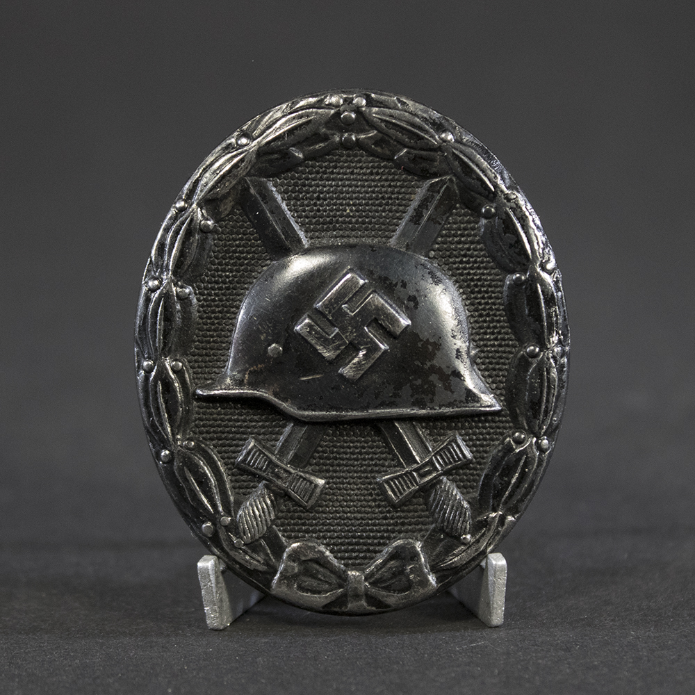 “Full view of a World War II German black wound badge, oval-shaped with a raised design featuring a helmet over crossed swords, surrounded by a laurel wreath.”