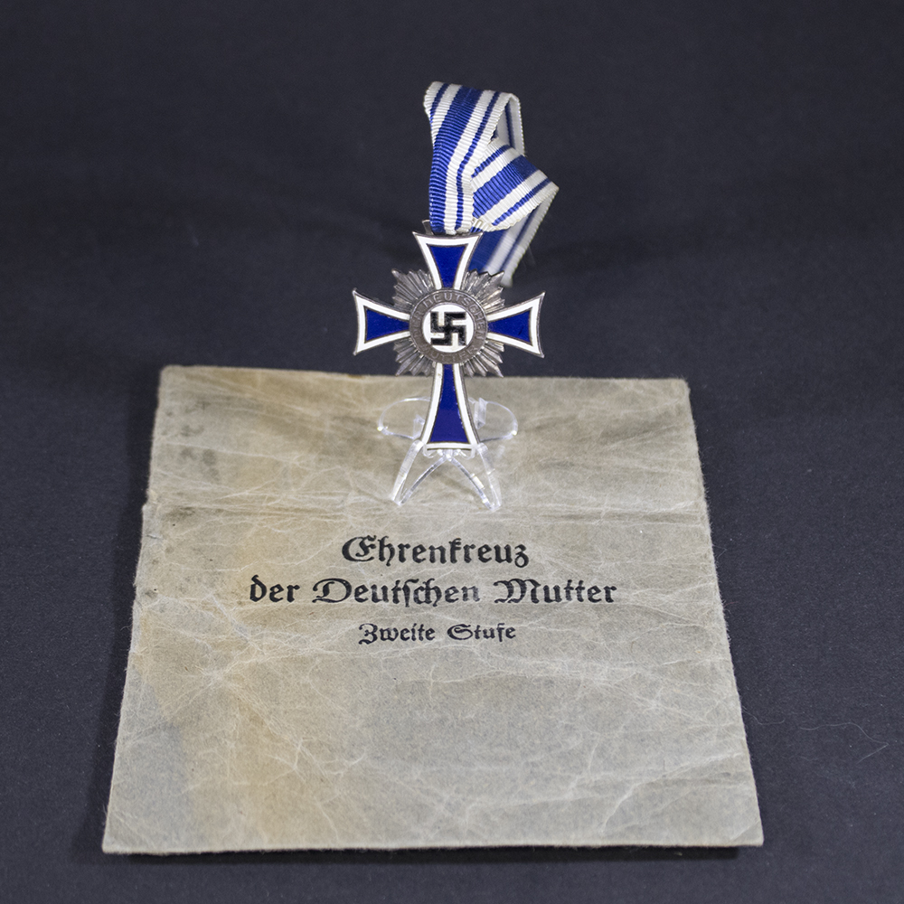 “Silver Mother’s Cross displayed alongside its original storage bag. The cross features a white and blue enamel design with a central swastika and inscription, while the bag is made of dark fabric with a flap closure.”