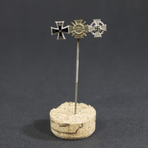 “Stick pin featuring miniature versions of three medals mounted side by side: the Iron Cross 2nd Class from World War I, the Frontkämpfer badge, and the 25 Years Faithful Service medal. The pin is metallic with detailed designs on each medal.”