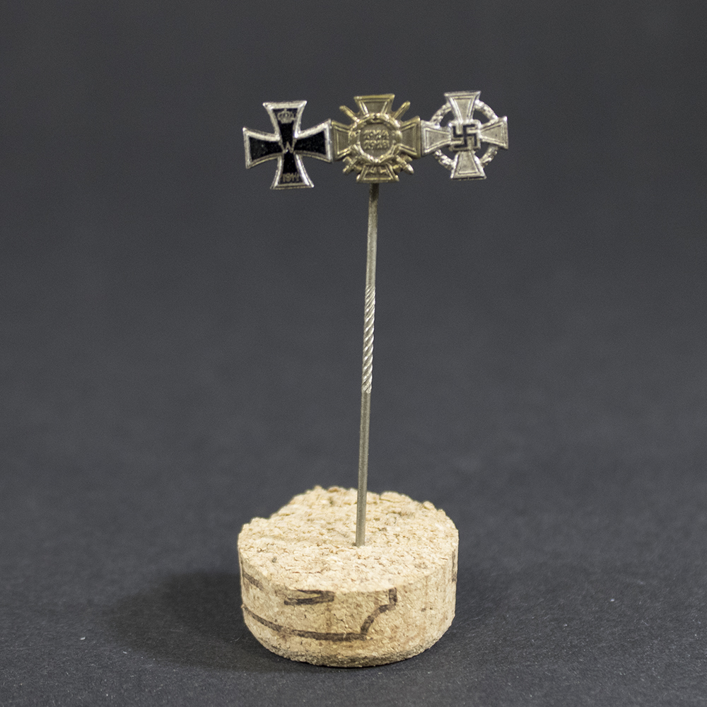 “Stick pin featuring miniature versions of three medals mounted side by side: the Iron Cross 2nd Class from World War I, the Frontkämpfer badge, and the 25 Years Faithful Service medal. The pin is metallic with detailed designs on each medal.”