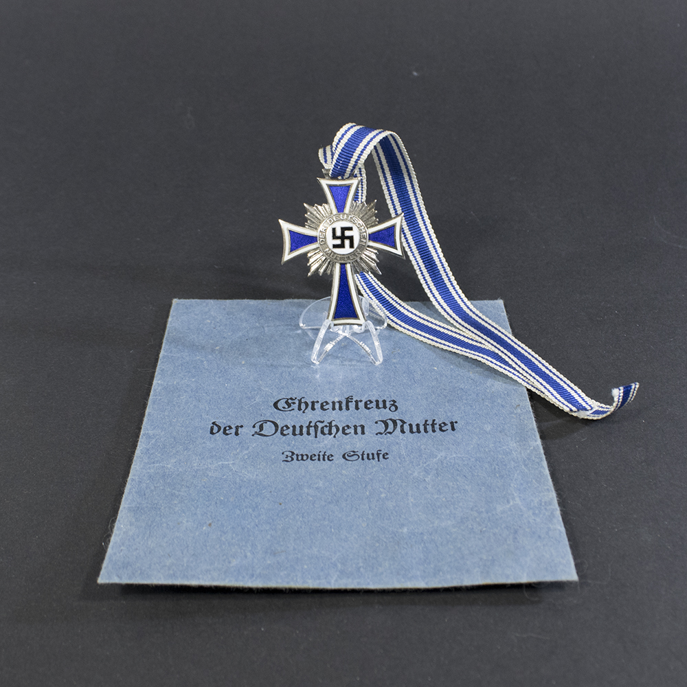 A World War II German Mother’s Cross medal displayed alongside its original presentation bag. The medal features a blue and white enamel cross with a central swastika and inscription, placed next to a textured blue bag.
