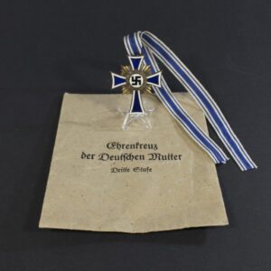 A World War II German Mother’s Cross medal displayed alongside its original presentation bag. The medal features a blue and white enamel cross with a central swastika and inscription, placed next to a textured brown bag.