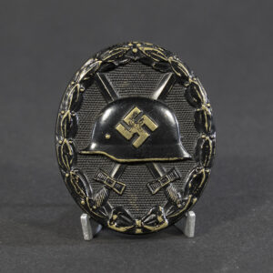 A World War II-era German black wound badge, oval-shaped with a raised design featuring a steel helmet over crossed swords, surrounded by an oak leaf wreath.