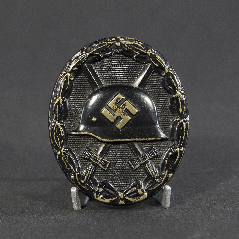A World War II-era German black wound badge, oval-shaped with a raised design featuring a steel helmet over crossed swords, surrounded by an oak leaf wreath.