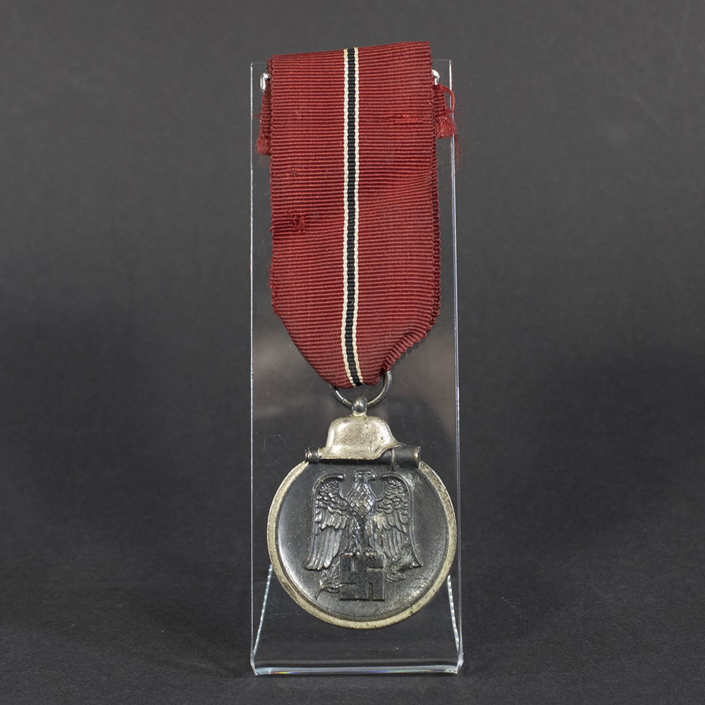 A World War II German Eastern Front Medal displayed in full view, showing its oval shape with a loop at the top for suspension. The medal features a darkened finish and a ribbon attached.