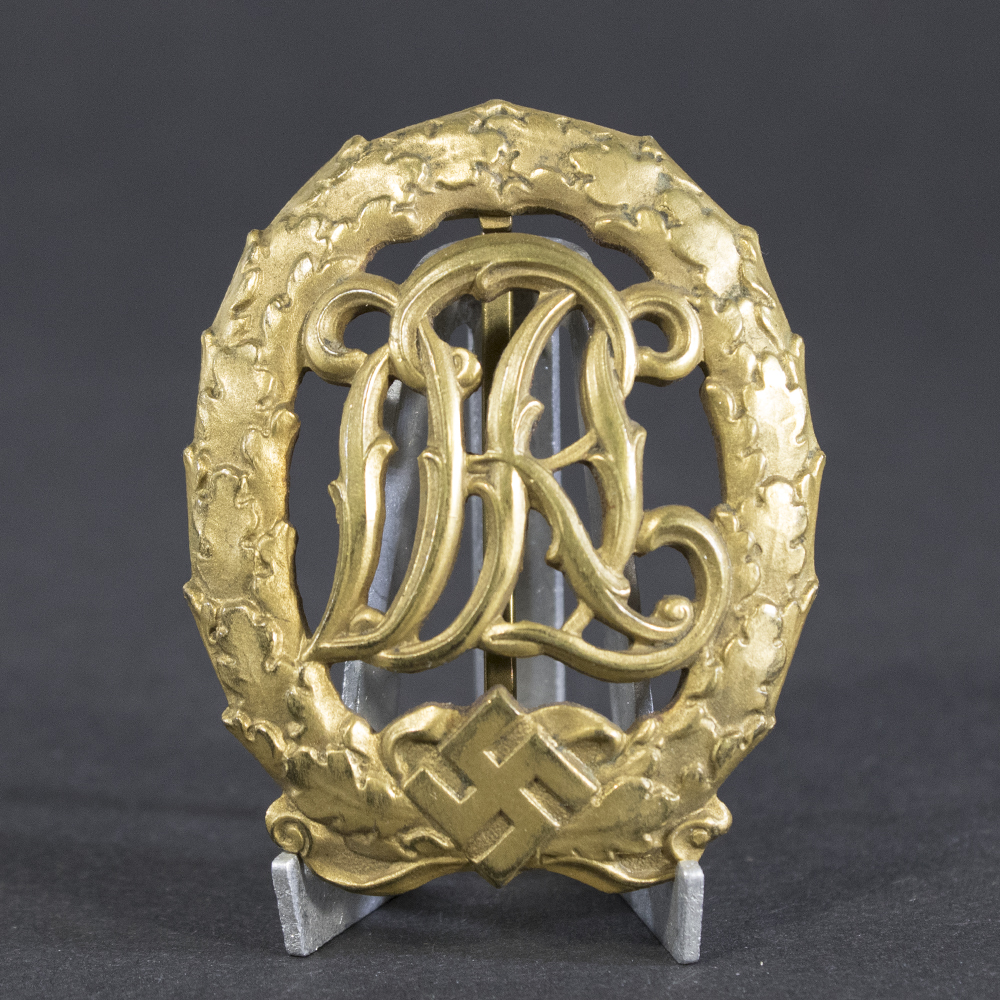 Front view of a World War II German golden DRL sports badge, featuring an ornate monogram of the letters ‘DRL’ surrounded by a laurel wreath with a gold finish.
