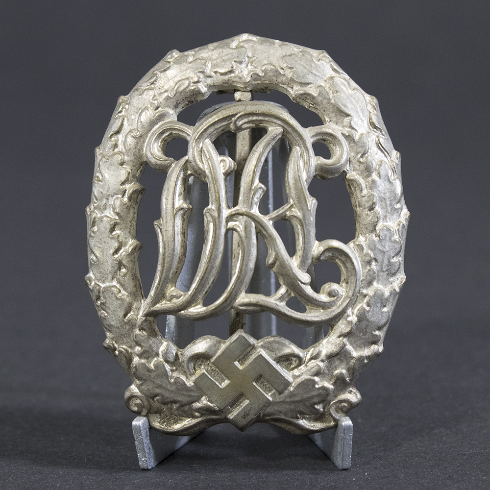Front view of a World War II German silver DRL sports badge, featuring an ornate monogram of the letters ‘DRL’ surrounded by a laurel wreath with a silver finish.