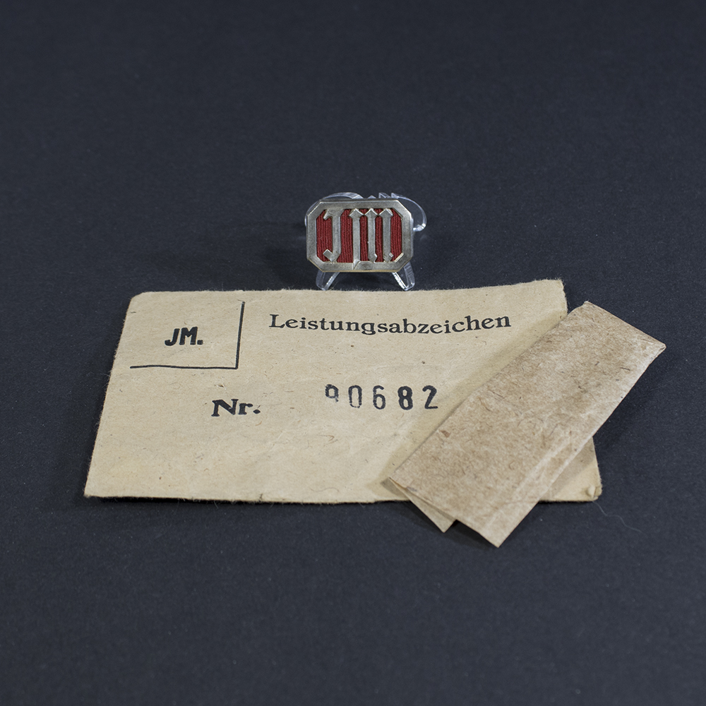 Full view of a World War II German Jungmädel membership pin displayed alongside its original paper bag.