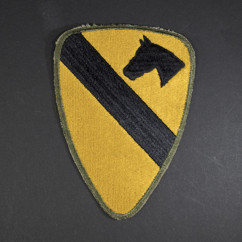 Front of WWII U.S. Army 1st Cavalry Division patch, featuring yellow shield with black diagonal stripe and horse head emblem.