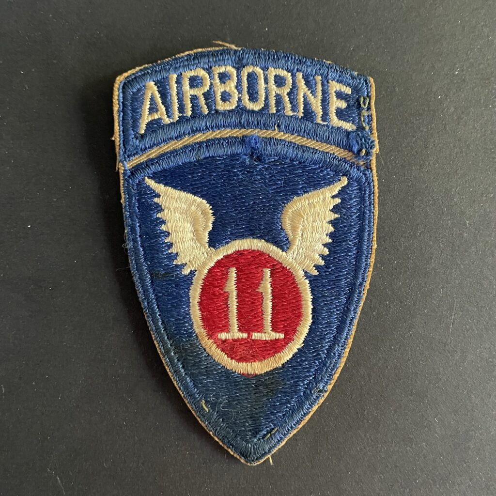 Front of WWII U.S. 11th Airborne Division patch, featuring blue shield with white winged parachute emblem and red border.