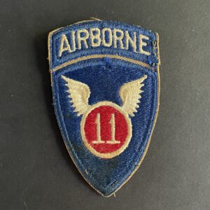 Front of WWII U.S. 11th Airborne Division patch, featuring blue shield with white winged parachute emblem and red border.