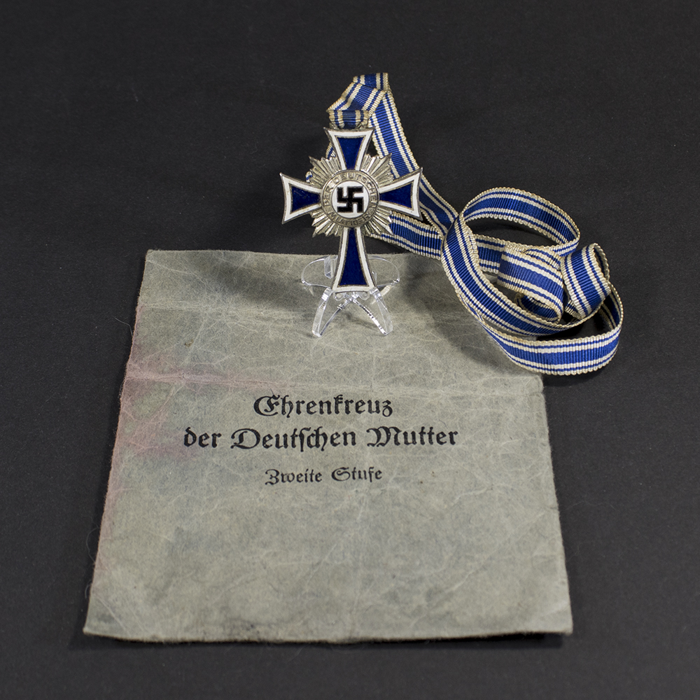 Silver-grade German Mother’s Cross from World War II displayed alongside its dark blue presentation bag, showing the full medal with ribbon and the bag lying flat.