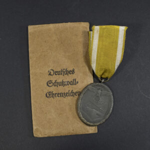 Full view of a World War II German Westwall Medal alongside its brown presentation bag, showing the complete medal with ribbon placed next to the labeled paper bag.