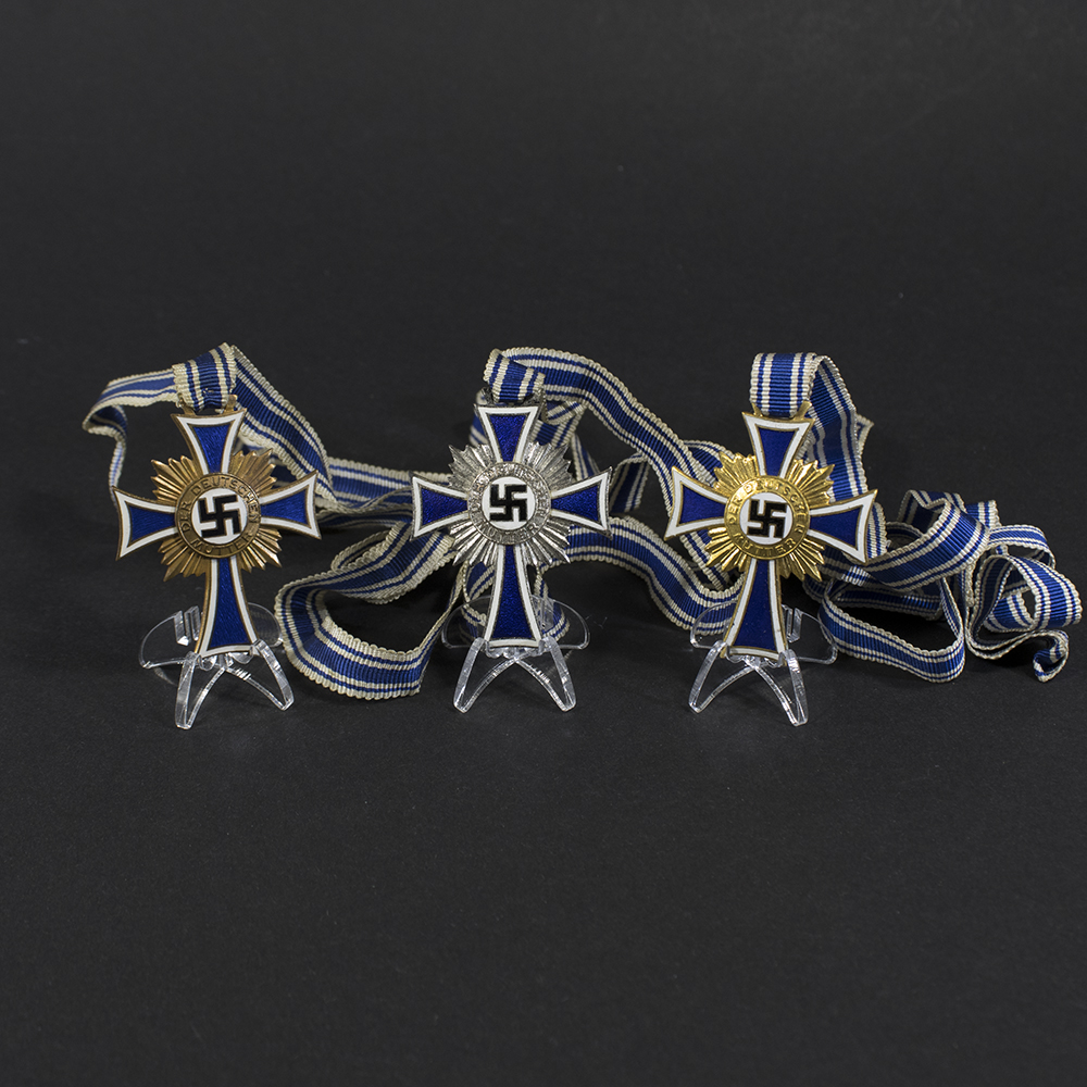 Overview photo of a complete Mother’s Cross set, showing the bronze, silver, and gold medals arranged side by side with their blue‑and‑white ribbon bows. All three crosses are visible from the front, displaying their enamel surfaces and central medallions.