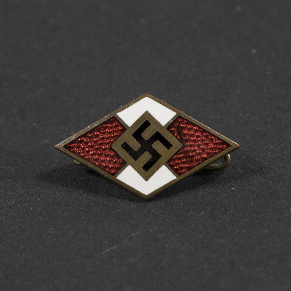 Overview photo of a WWII Hitler Youth membership pin, showing the small diamond‑shaped badge with its red‑white‑black enamel design. The pin is displayed from the front with its metal fastening visible behind it.