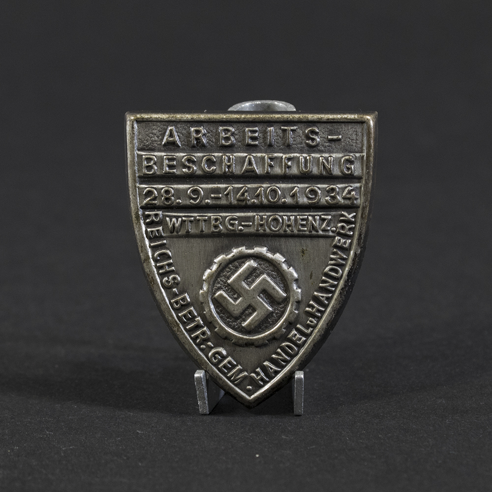 Overview photo of a WWII German DAF “Arbeitsbeschaffung” tinnie, showing the full badge with its circular shape, raised relief design, and metal finish. The front inscription and emblem are visible, along with the overall condition of the piece.