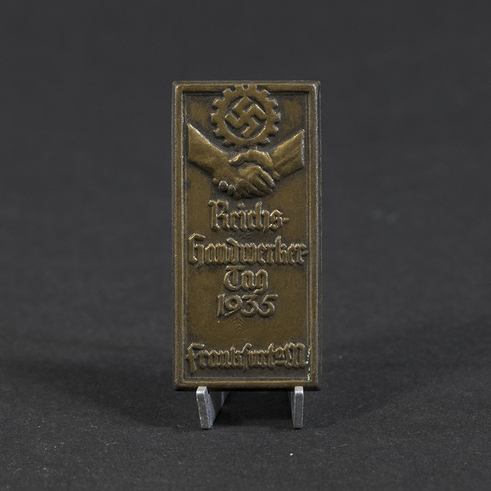 Overview photo of a 1935 “Reichshandwerkertag Frankfurt a. M.” tinnie, showing the full badge with its rectangular shape, raised relief design, and metal finish. The event title and central emblem are visible, along with the overall condition of the piece.