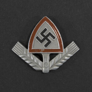 Front view of a WWII German RAD cap badge, showing the metal emblem with its stylized spade shape, central wheat‑sheaf motif, and surrounding raised details. The contours, texture, and metallic finish of the badge are clearly visible.