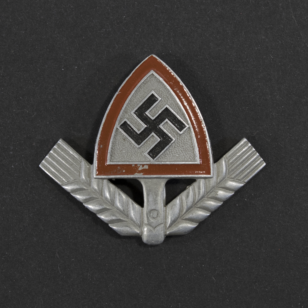 Front view of a WWII German RAD cap badge, showing the metal emblem with its stylized spade shape, central wheat‑sheaf motif, and surrounding raised details. The contours, texture, and metallic finish of the badge are clearly visible.