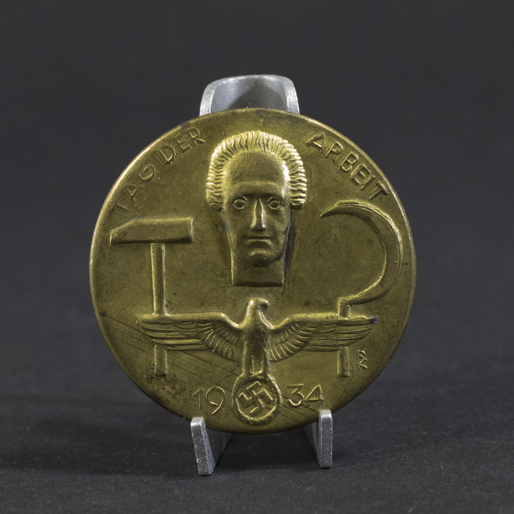 Overview photo of a WWII German “Tag der Arbeit 1934” tinnie, showing the full badge with its raised relief design, event inscription, and metal finish. The overall shape, imagery, and condition of the piece are clearly visible.