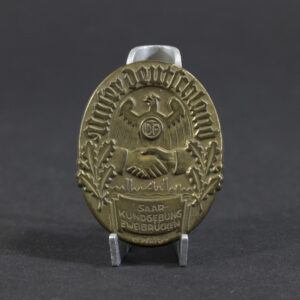 Overview photo of a WWII German “Unser Deutschland – Saar Kundgebung Zweibrücken, 6. Mai 1934” tinnie, showing the full badge with its rectangular shape, raised relief design, event inscription, and metal finish. The overall layout, imagery, and condition are clearly visible.