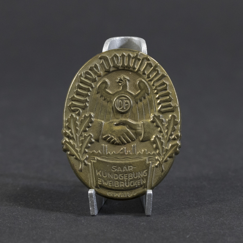 Overview photo of a WWII German “Unser Deutschland – Saar Kundgebung Zweibrücken, 6. Mai 1934” tinnie, showing the full badge with its rectangular shape, raised relief design, event inscription, and metal finish. The overall layout, imagery, and condition are clearly visible.