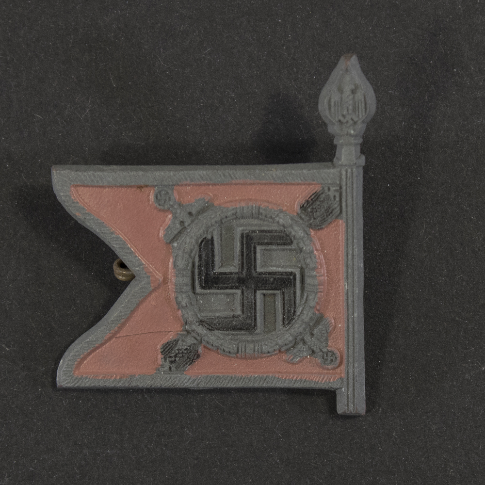 Close‑up of the front side of a small plastic Winterhilfswerk miniature flag associated with the Führerbegleitbataillon. The rectangular flag shows an eagle emblem and raised lettering molded into the plastic. The surface has a glossy finish, and the relief details and edges are clearly visible despite minor wear.