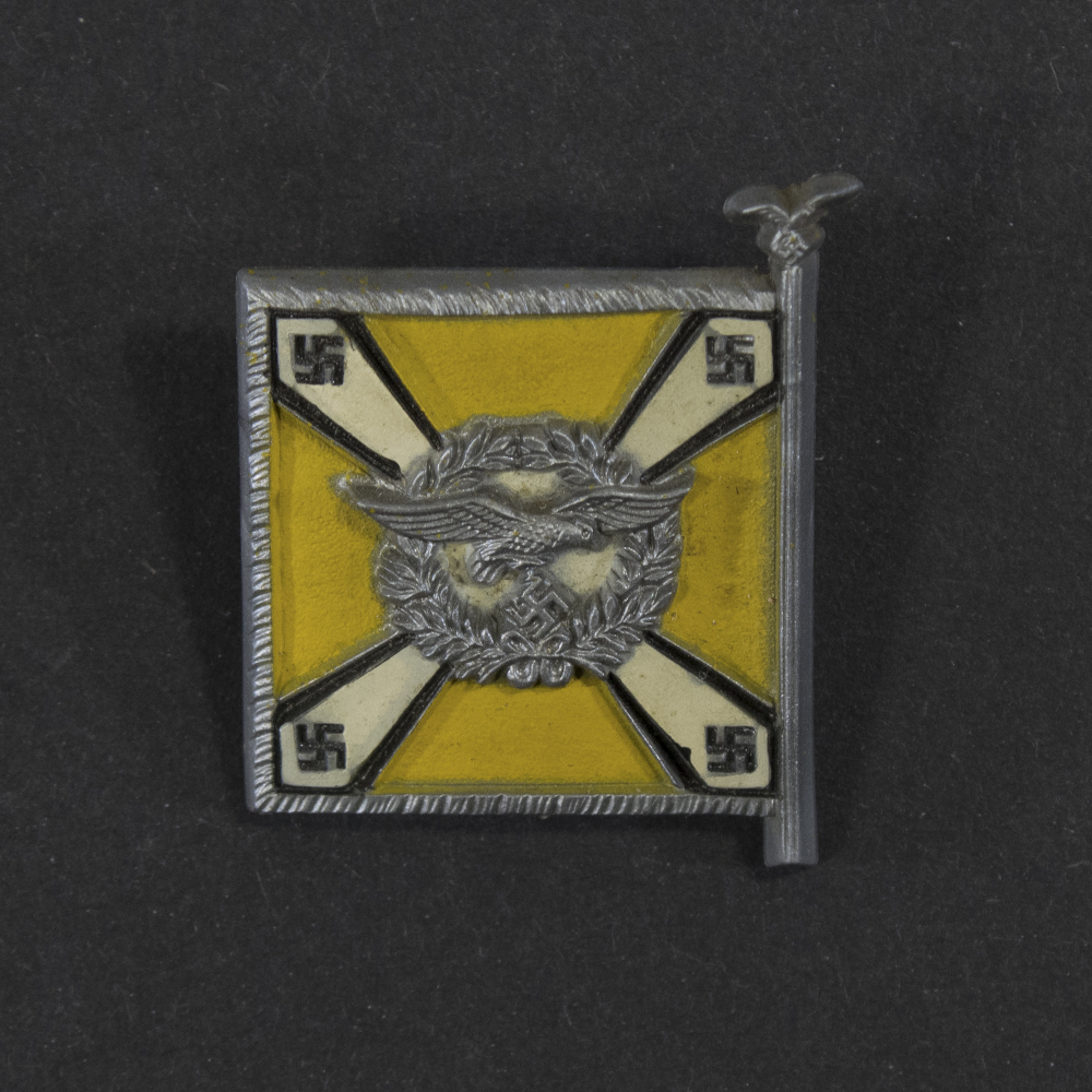 Close‑up of the front side of a small plastic Winterhilfswerk miniature flag associated with the Fliegertruppen. The rectangular flag shows an eagle emblem and raised lettering molded into the plastic. The surface has a glossy finish, and the relief details and edges are clearly visible despite minor wear.