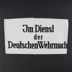 Overview of a World War II-era German cloth armband, white fabric with black lettering reading “Im Dienst der Deutschen Wehrmacht,” displayed flat against a neutral background.
