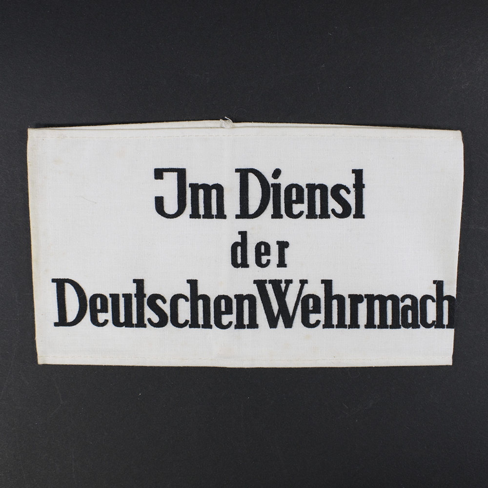 Overview of a World War II-era German cloth armband, white fabric with black lettering reading “Im Dienst der Deutschen Wehrmacht,” displayed flat against a neutral background.