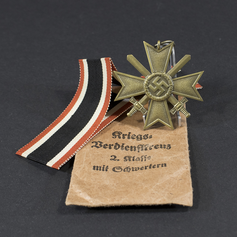 Overview of a World War II-era German War Merit Cross 2nd Class medal set displayed with its original paper issue bag, showing the bronze cross and accompanying packaging on a neutral background.