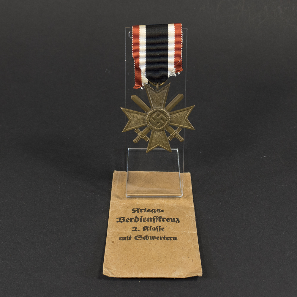 Overview of a World War II-era German War Merit Cross 2nd Class medal set displayed with its original paper issue bag, showing the bronze cross and accompanying packaging on a neutral background.
