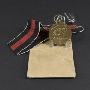 Overview of a World War II-era German Sudetenland Occupation Medal displayed together with its original paper issue bag, showing the medal suspended from its ribbon alongside the packaging on a neutral background.