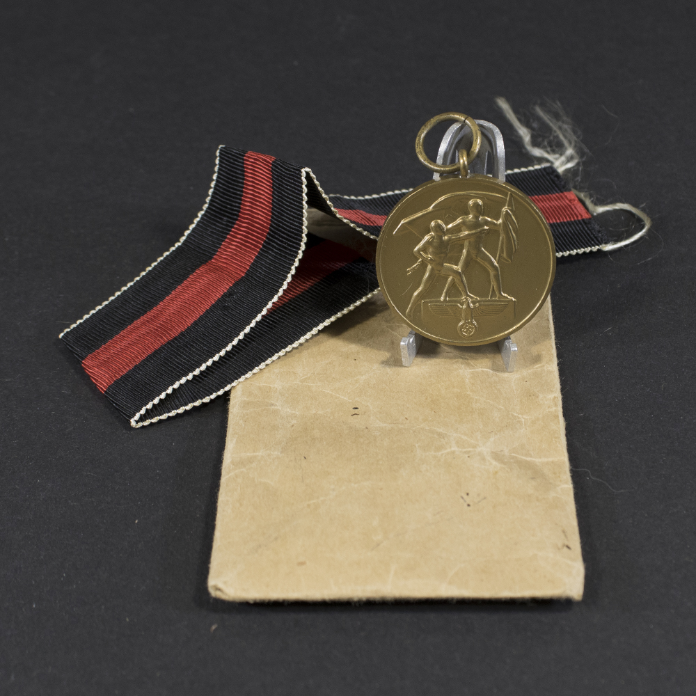 Overview of a World War II-era German Sudetenland Occupation Medal displayed together with its original paper issue bag, showing the medal suspended from its ribbon alongside the packaging on a neutral background.