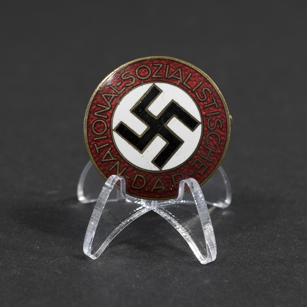 Overview of a World War II-era German NSDAP membership pin displayed on a neutral background, showing its circular enamel design with red, white, and black colors.
