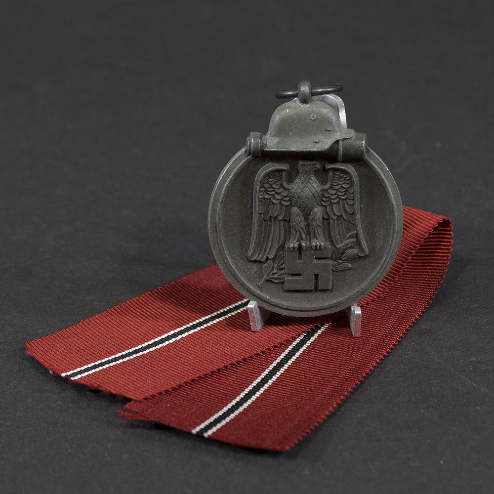 Overview of a World War II-era German Eastern Front Medal (Ostmedaille), shown with its ribbon, displaying the round medal suspended from a red, white, and black striped ribbon against a neutral background.