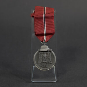 Full view of a WWII German Eastern Front Medal (Ostmedaille) suspended from its red, black, and white ribbon. The circular medal features a military-style design with a helmet and stick grenade motif. The metal shows age-related wear and patina. The suspension ring is maker-marked “19,” identifying the manufacturer as Friedrich Orth.