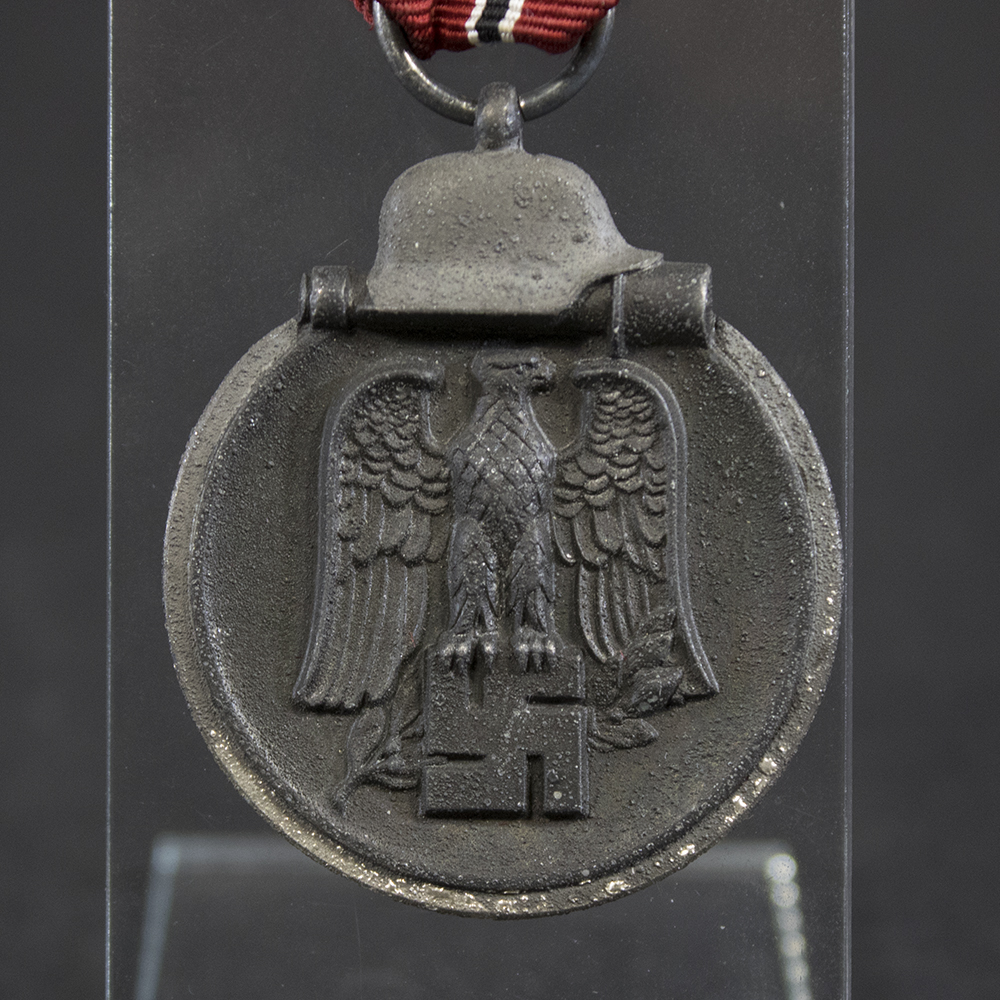 Close-up of the front of a WWII German Eastern Front Medal. Visible details include the national emblem above a military helmet resting over a stick grenade. The raised design remains well defined, with moderate wear and darkened finish from age.